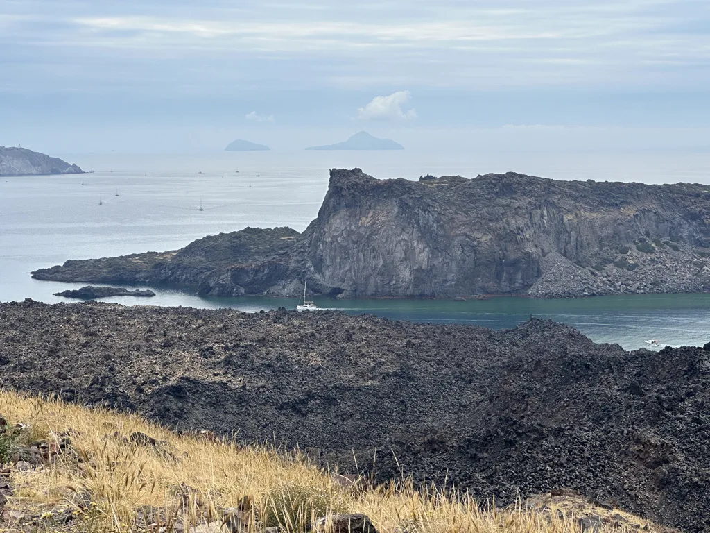 View across volcanic rock to a small bay with a sailboat, distant islands on the horizon