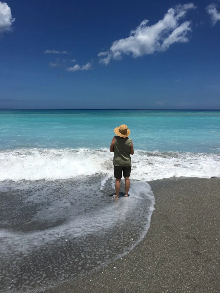 Person in straw hat standing at the edge of a turquoise sea, foam swirling around their feet