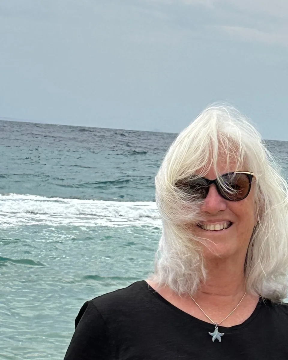 M. Joy Gorence on a beach in front of the sea, smiling, white hair tossed by the wind